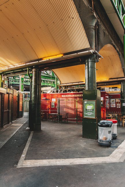 This image depicts a vibrant outdoor area near Borough Market, featuring a modern, partially covered space with a corrugated beige roof supported by black metal columns. The ground is paved with smoot